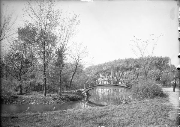 View from path of a footbridge over the lagoon in Tenney Park. A man is walking along the path on the right. Trees are along the shoreline.