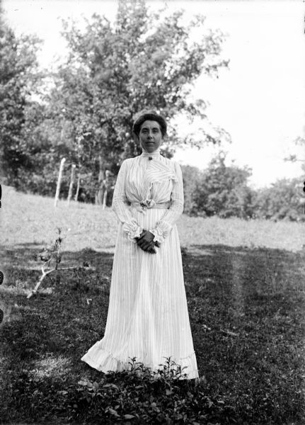 Full-length outdoor portrait of Anna McConnell standing in the shade in a field, with trees in the background.