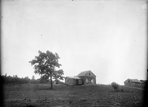 View across field of a deserted cabin at Camp Douglas. Another group of cabins are behind a fence on the right. Trees and foliage are in the background.