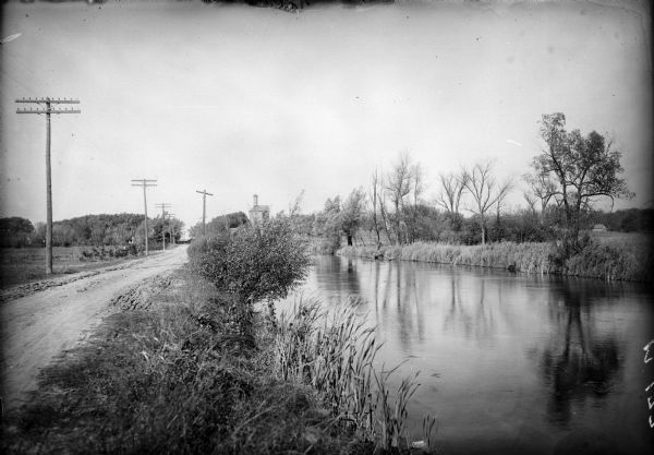 View along the Catfish River, with shrubs and grasses covering the riverbank. On the far left are electrical poles along a dirt road bordering a field that stretches to the horizon. In the far background is a tall industrial building with a chimney among trees.
