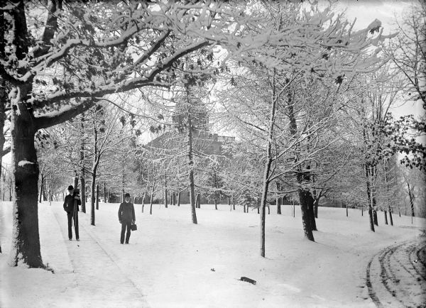 Winter scene near Main Hall (Bascom Hall) drive on the University of Wisconsin campus. View looking uphill towards two men standing on and near a sidewalk. Both men are carrying cameras on tripods. Trees are on the snowy lawn, with Main Hall in the background.