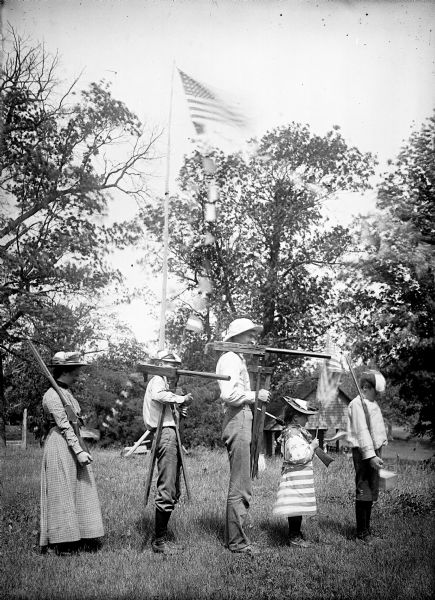 View from side of a Fourth of July Procession of five people standing in a line carrying various types of weapons and dressed in festive clothing. There is a flag flying from a tall pole in the background, with a number of smaller flags or banners flying below.