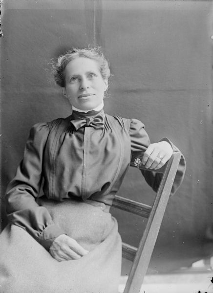 Portrait of Jessie Turvill Thwaites, wife of the Wisconsin Historical Society Director Ruben Gold Thwaites. She is sitting sideways in a chair, and is holding a flower in one hand resting on the back of the chair, with the other hand is resting in her lap. A dark backdrop is in the background.