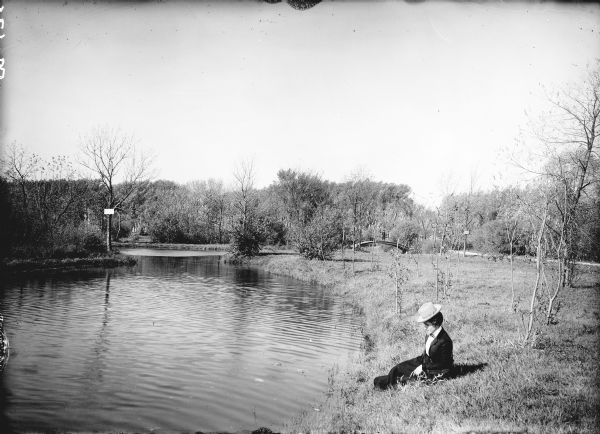 Original caption reads, "A scene in Tenney Park." View along shoreline towards a woman sitting in the grass along the shore of the lagoon. She is wearing a hat and is leaning on a handbag or camera case. In the background behind her a man stands on a footbridge in the background. To the right of the footbridge is a wide path.