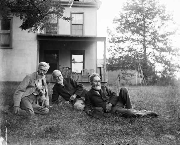 Outdoor group portrait of the Turvill brothers. All three men have beards. They are posing sitting on the ground in front of the family home. The man on the left is posing with a small dog.