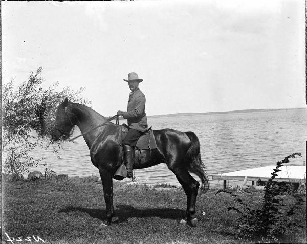 Outdoor portrait of Mr. Olin sitting on a horse on a lawn near the shoreline of a lake. The far shoreline is in the background. Near the shoreline on the right is what appears to be a dock, or the roof of a boathouse.