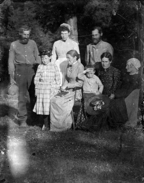 Outdoor group portrait of eight members of the Thwaites family. Three of the women are sitting, and one has a young boy sitting in her lap. Two men, a woman, and a young girl stand.