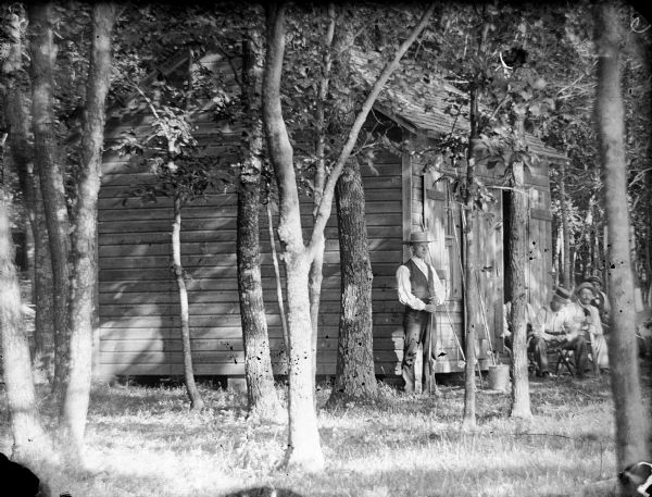 View through trees towards a cabin. One man holding a rifle stands in the center at the left front side of the cabin, and a group of four men sit on the right near the open doorway. Fishing poles and a bucket are in front of the cabin window.