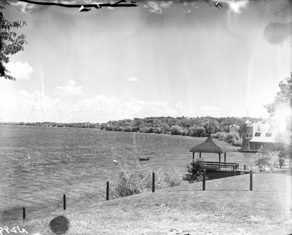 View from sloped lawn of the Lake Mendota shoreline, with the Olin boathouse in the foreground, and the University of Wisconsin-Madison boathouse on the right. A boat just off the shoreline is anchored to a floating barrel. In the distance is the City Boathouse, designed by Frank Lloyd Wright and built for the city of Madison.
