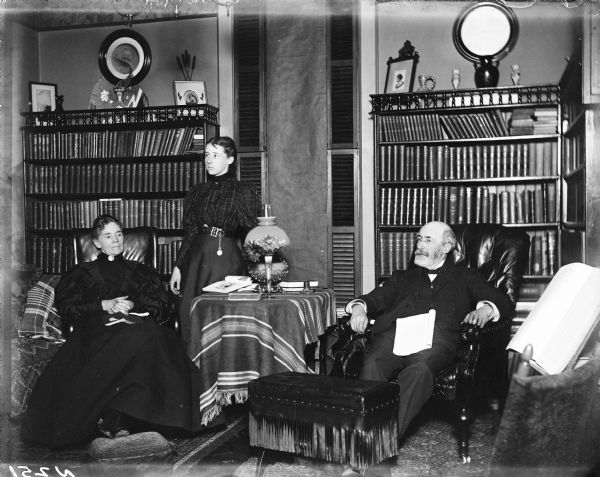 A Morris family group portrait in their home library. Full bookcases surround a window. Mr. and Mrs. Morris are seated, while their daughter stands between them. A table with a lamp is in the middle of the two chairs. Candlesticks, vases, framed pictures and a mirror stand atop the bookcases.