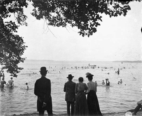 Original title reads, "Bathers assemble [swimmers in Lake Monona]." Women and men, wearing suits, hats, and long dresses, are standing near the lake shoreline looking on as others swim in the lake.