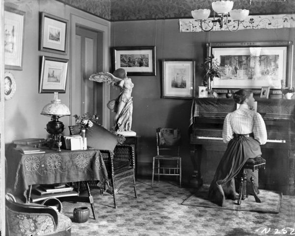 Interior view of the William A.P. Morris residence parlor. A young woman is sitting at a piano on the far wall, looking over her shoulder to her right. Framed pictures hang on the walls, and there is a large sculpture on a table near a doorway in the left corner. Two chairs and a table are along the left wall.