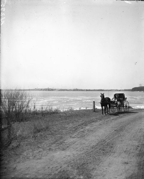 View down dirt road towards Madison from Elmside. There is a horse and buggy on the side of the road near the shoreline, with a woman wearing a veil sitting inside. The buggy top is folded halfway down. Houses line the far shoreline, and the Wisconsin State Capitol is on the left.