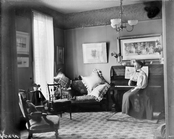An interior view of the parlor of the William A.P. Morris residence. On the right is a young woman sitting sideways in front of a piano looking towards the window on the left. She is wearing a shirt with puffy sleeves, a long skirt, and a bow at her neck, with her hair pulled back. Framed art work hangs on the walls. In front of the window is a sofa with four large pillows, and two chairs.