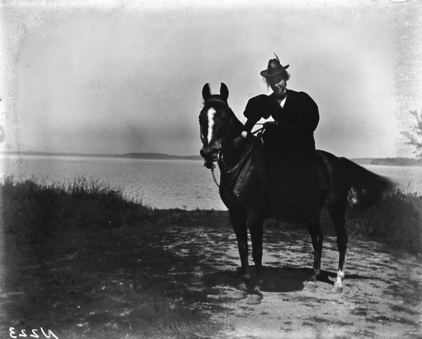 Outdoor portrait of Elsie Stevens sitting on "Kittie" the horse. They are posed with the lake in the background.
