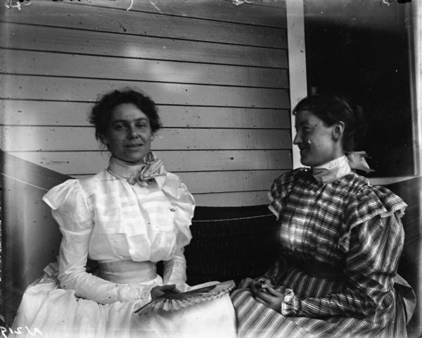 Outdoor portrait of Miss Oakle and Richardson sitting together in a hammock on a porch. Both women hold hand fans and wear dresses with puffy sleeves and high collars.
