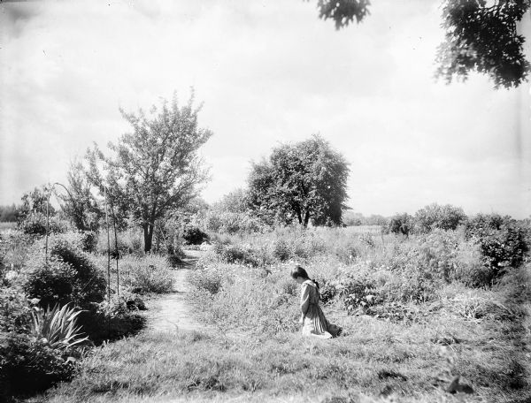 A unknown girl is kneeling in Turvill garden. She is wearing a dress and her hair is braided.  There is a path in the center and the gardens surround her. Trees are in the background.