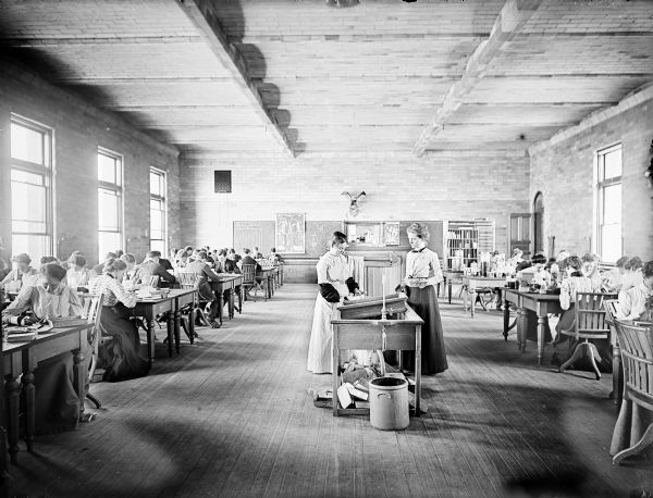 University of Wisconsin-Madison biological laboratory. Two women are standing in the center of the room at a sink, and men and women sit at tables on the right and left near large windows.