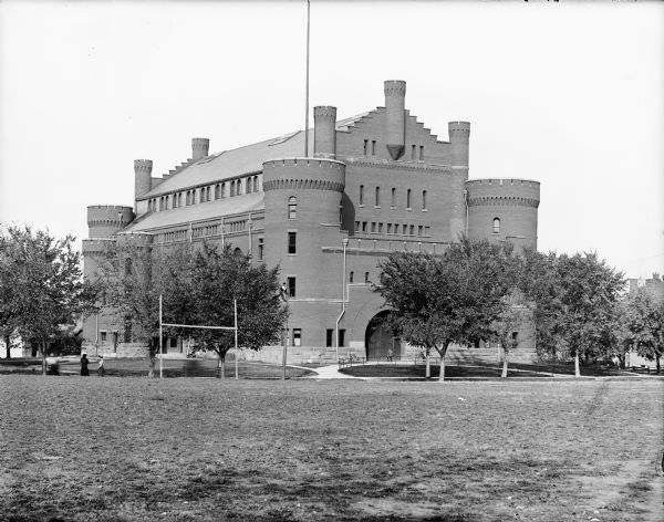 View across lawn of the University of Wisconsin Armory building (Red Gym). A man is sitting on a railing in front of the gym entrance door. Another man is working high up on a wooden pole in front of the Armory along Langdon Street.