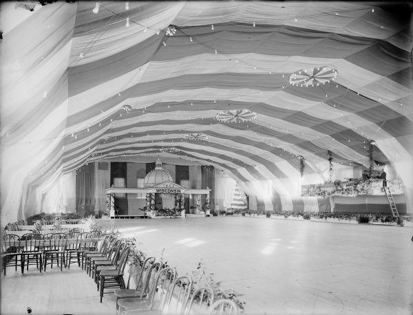 The University of Wisconsin Gymnasium decorated for the junior prom. Lights, fabric drapes, and foliage are strung along the ceiling and walls. Chairs, plants in boxes along a railing, and tables are positioned about the room. The stage is constructed in the form of a pavilion with a dome, with a "Wisconsin" sign centered above columns under the date "1903." Men are standing in a raised balcony on the right side.
