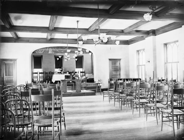 Interior view of Cornelia Vilas Guild Hall in the Women's Club on the University of Wisconsin-Madison campus. The stage is set with chairs in front of three windows, and a table set with a flower arrangement, punch bowl, and other tableware. In front of the stage on a lower platform are two chairs and a desk, and a small table with a pitcher of water and two glasses. An aisle between rows of chairs leads to the stage.