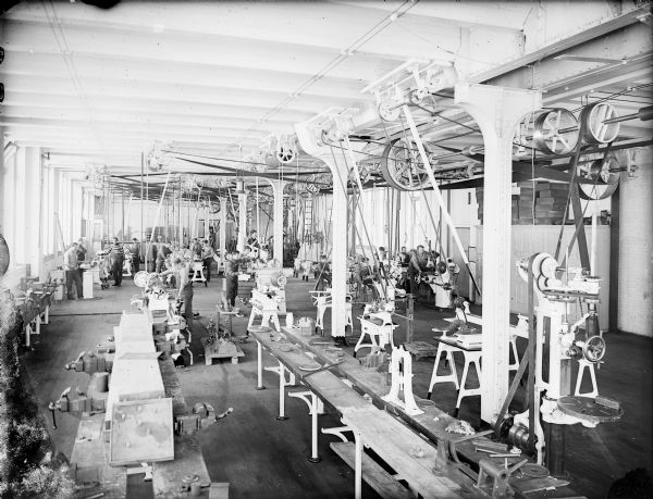 Elevated view of the University of Wisconsin-Madison machine shop. A large group of men are working various machines in the workshop area. Long wooden work benches are in the right foreground.