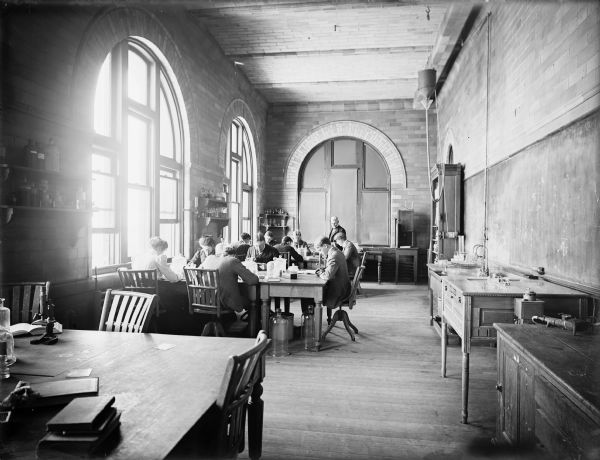 Interior view of a histological laboratory at the University of Wisconsin-Madison. A group of students, men and women, are sitting around two tables in front of two large arched windows on the left side. A man wearing a suit stands in the back of the room in front of another arched window with the shades pulled. Cabinets, equipment and sinks line the right side of the room.