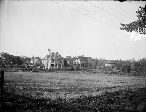 View across mowed field of two University of Wisconsin professors residences. On the left is the residence of Professor Harry L. Russell and to the right is the residence of Professor Franklin H. King. There are windmills in the neighborhood. A round barn is on the far right.