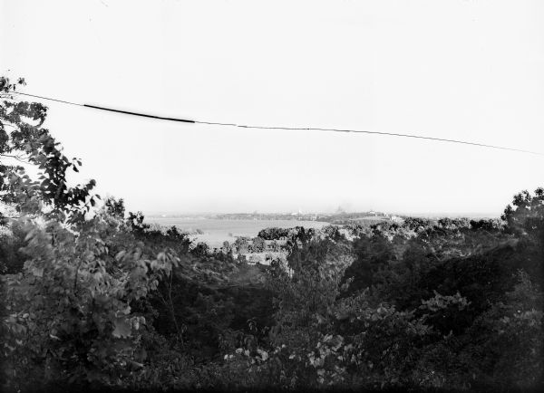 Elevated view of Madison through trees. Lake Mendota, buildings of the University of Wisconsin-Madison, including the Washburn Observatory are along the shoreline on the right. The Wisconsin State Capitol, church buildings, and a water tower rise above the trees on the far shoreline.