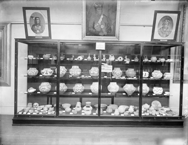 The Wisconsin Historical Society Museum display of Cliff Dweller Pottery from the Robert Laird McCormick Collection. Pottery is displayed in a glass case with three paintings hanging above. The paintings are titled, from left to right: "Esense," "Souligny" and "Keokuk."