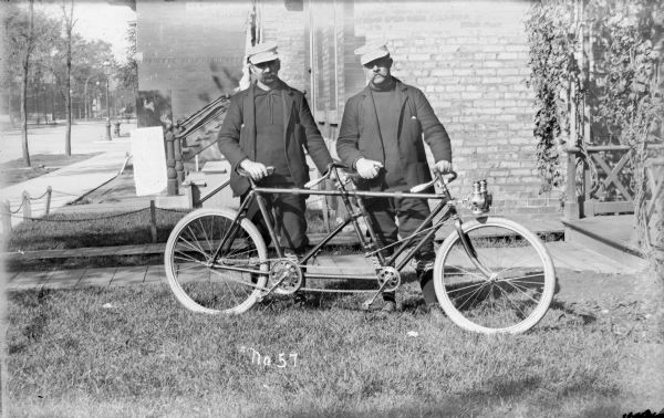 An outdoor portrait of two men with a tandem bicycle. The men stand in a yard near a front walk leading up to a porch. In back of the men is the side of a brick building, with a sidewalk and street on the left. The men wear hats, sweaters, coats, stockings, button-up shoes and knickerbockers.