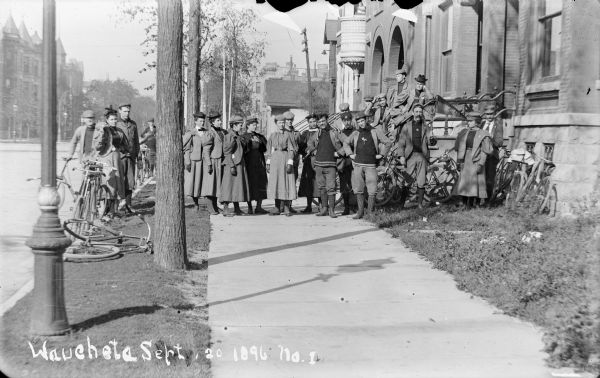 View down sidewalk of a group of men and women standing near a large brick building. Bicycles are leaning against the curb and the side of the building. The women are wearing dresses, neckties, button-up shoes and dark gloves. The men wear sweaters, jackets, button-up shoes and knickerbockers.