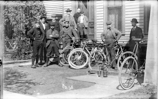 Group of men, and a young boy, standing in the small front yard of a house at a bicycling outing. A woman stands in an open doorway at the top of the steps. A man on the far right wears a suit and appears to be holding a cigar. Bicycles are leaning against a railing in front of the house. The men wear sweaters, jackets, hats, stockings, button-up shoes and knickerbockers. On top of a wood sidewalk in the yard is what appears to be a hand-pump with a long handle for putting air into bicycle tires.

