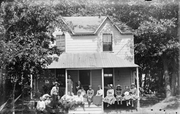 Group portrait of men and women at a gathering at Jolly Lodge. The men are wearing knickerbockers, and the women are wearing long skirts with puffy sleeved blouses. They are posing on the front porch of a home in the woods, and one of the women is holding a banjo. On the porch behind the group, one woman is standing near a column, and one woman is sitting in a chair, with a dog sitting in front of them. There is a bicycle pump hanging on the right porch wall, and bicycles are leaning against the wall on the left side of the porch. Hammocks are tied to trees on the far left.