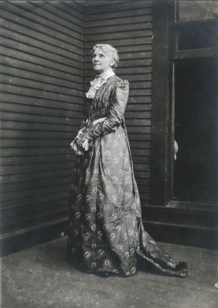 Full-length portrait of Blanchard Harper, who is wearing a long dress with lace collar and cuffs. She appears to be standing in the corner of a porch, with a window behind her on the right.