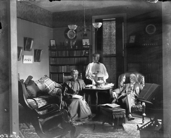 Group portrait of the Morris family in their family library. Mr. and Mrs. Morris sit in two chairs, while their daughter stands in between them behind a table. Bookshelves filled with books are in the background.