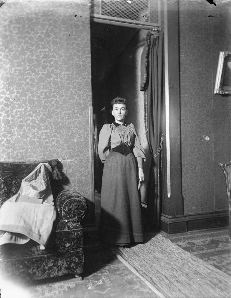 Full-length portrait of an unknown women standing in a doorway. The tall doorway has a drape on a pole just underneath a transom window or screen. A coat is draped on a couch on the left, with a book resting on top of it. The edge of a chair is on the right near a framed painting on the wall.