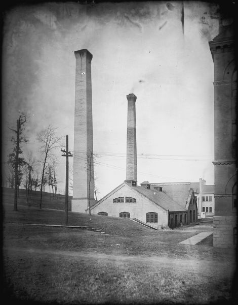 University of Wisconsin heating plant, with two tall chimney stacks. Electric lines are in the foreground. There are trees on a hill on the left. On the right is the side of Science Hall.