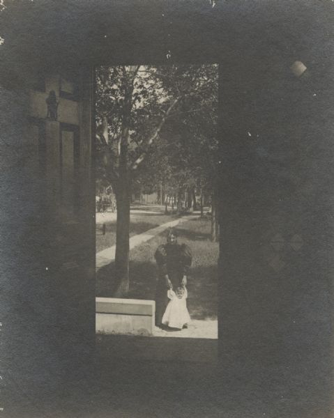 View through open doorway, looking out towards Mrs. Lucius Fairchild and her granddaughter, Frances Bull, standing on a sidewalk. Original caption reads, "Frances Fairchild  Bacon Gary (1895-1976) wearing mourning clothes. Fairchild Mansion over Lake Monona, former home of S.C. Fairchild. Trees, a sidewalk and a horse-drawn wagon are in the background.