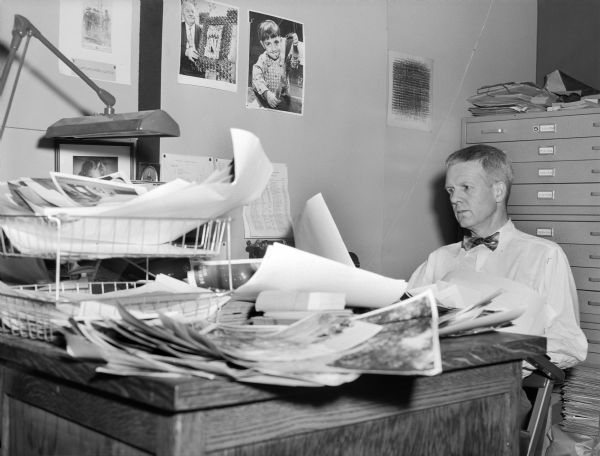 Paul Vanderbilt, Curator of the Iconographic Collections (1954-1972), State Historical Society of Wisconsin, examining photographs received from the <i>Milwaukee Journal.</i> His desk is covered with piles of photographs and several prints are tacked to the wall. Flat files are behind him.