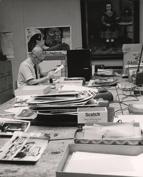 Paul Vanderbilt works at a table in the Iconography Section at the Wisconsin Historical Society. Stacks of photographs and boxes cover the table, along with tape dispensers and other office supplies. A poster, and a painting of Nellie Jane Manning, are hanging on the back wall. Filing cabinets are on the left.