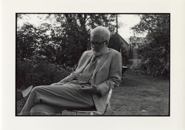 Paul Vanderbilt sitting in a painted metal chair in George Talbot's (Vanderbilt's successor) backyard on Jenifer Street. He is wearing a plaid jacket, trousers, patterned necktie and eyeglasses. Foliage, trees and dwellings are in the background.