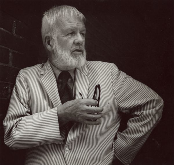 Paul Vanderbilt posed against a dark background. He has his left hand on his hip, and in his right hand he holds his glasses at chest level. He is wearing a striped jacket and dark necktie and has a beard and moustache.