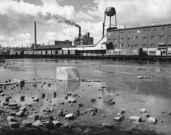 A diagonal view down the canal of the Menasha Woodenware Company. Along the opposite side of the canal are boxcars on an elevated railroad track over the water. In the foreground are rocks and old pier supports. In the background are industrial buildings, a water tower and smokestacks releasing smoke into the air.