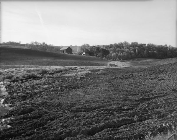 View across plowed field towards barn and farm buildings along CTH KP in the early morning. In the background are rolling hills and a ridge covered with trees.