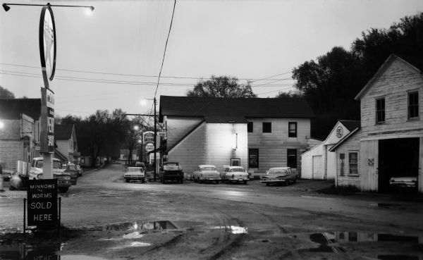 Texaco gas station on the left and cars parked on the road next to buildings in downtown Beetown. The road is rutted and muddy with puddles. Several signs can be seen, Texaco Perry Gas, Minnows and Worms Sold, Cities Service, Special Holiday Beer, Bob's Tavern, Ice, 7-up, V-C Fertilizers and Orange Crush. The truck door advertises Beetown [???] & Hardware.