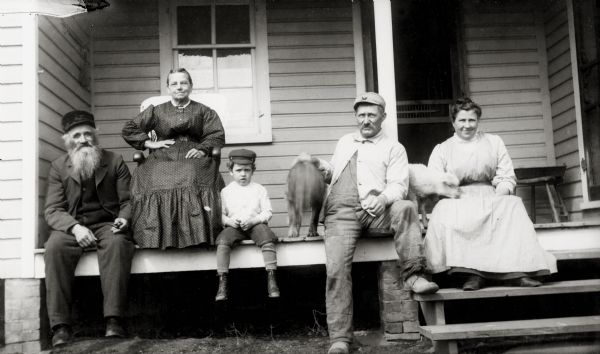 Two men and a young boy sit on the edge of the porch. The man on the left is wearing a suit and hat, the boy and the other man are wearing work clothes and caps. Two women, one in a chair and the other on the porch steps, are wearing dresses. The man on the right has his hand on the back of a dog that has his tail (wagging) towards the camera. A lamb is between the man and woman on the right.