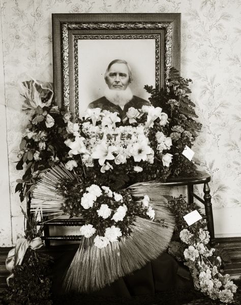 Funeral floral arrangement for William Jaeger. His framed portrait sits on two wooden chairs. He is wearing a suit and has a neckbeard. Flower arrangements are positioned on the chairs and on the floor in front of the portrait. The center floral arrangement has "Vater" spelled out on top of the flowers.