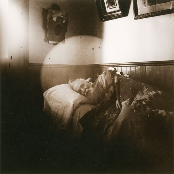 An elderly man is lying on his side, against the corner of a wall, facing the camera, with a pillow and quilt. Wainscoting covers the wall behind him, and two framed photos and a paper holder hang on the wall above.
