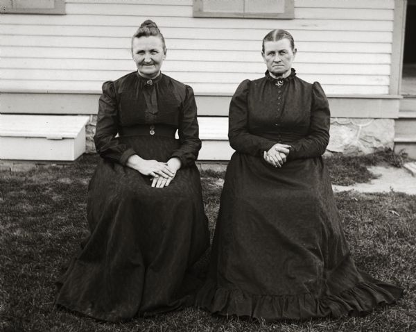 Two women are sitting in chairs on the lawn in front of a house. They are wearing dark dresses.